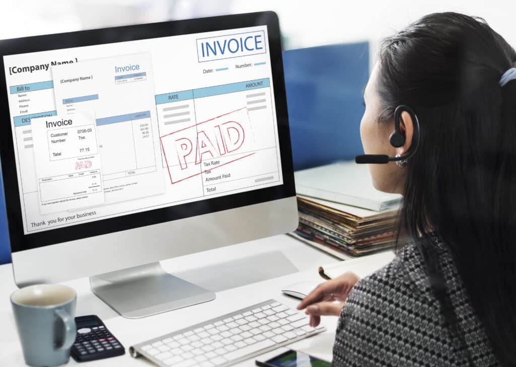 A woman wearing a headset with a microphone intently looking at a computer monitor displaying an invoice example with a red 'PAID' stamp.