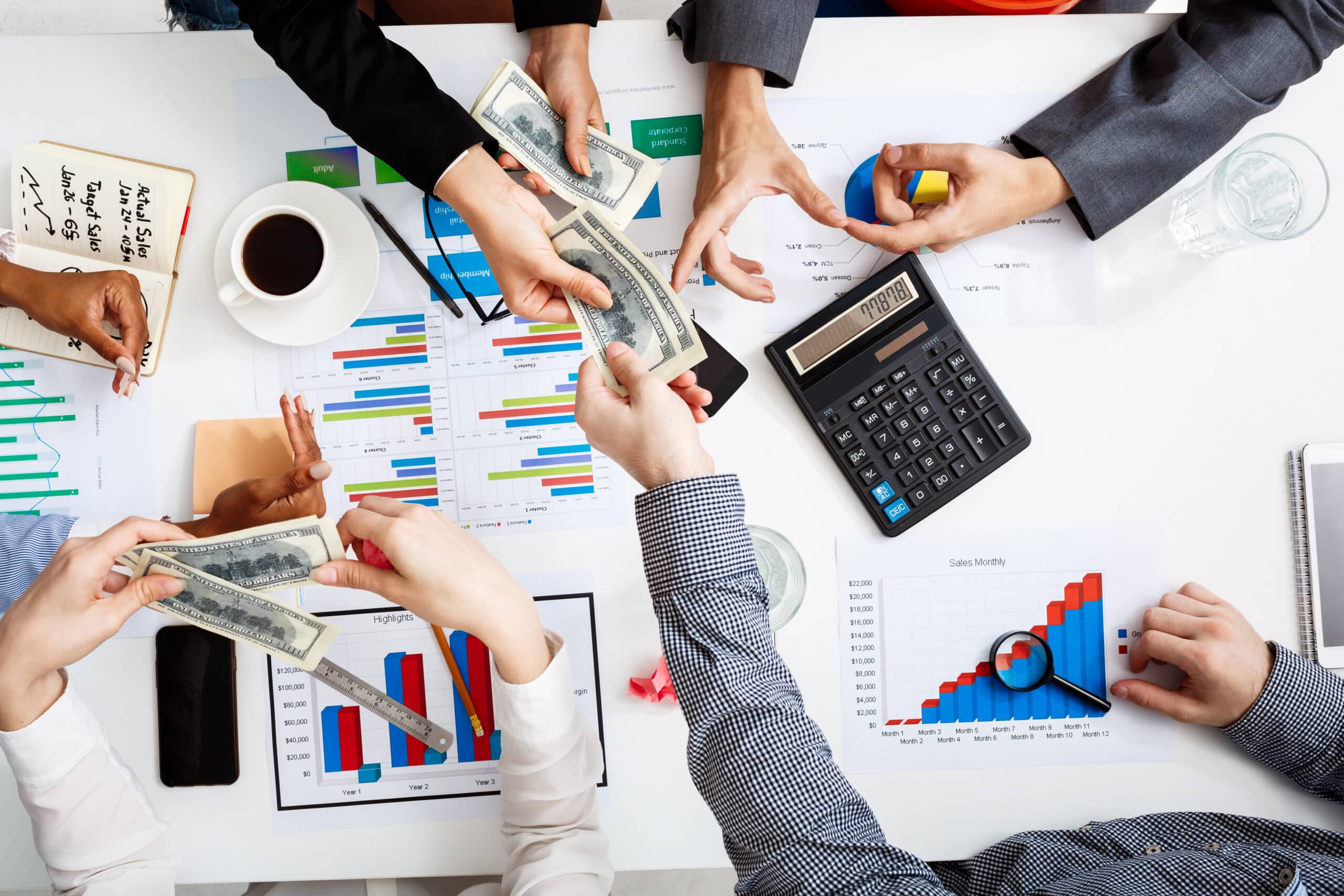 Beberapa tangan sedang bertransaksi uang di atas meja dengan grafik keuangan, kalkulator, dan kopi, menggambarkan pengelolaan keuangan bisnis online.